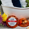 Two Citrus Rosemary Shower Steamers from The Gifted Hemp, placed in front of a bath setup with a towels and flowers, wrapped in orange foil