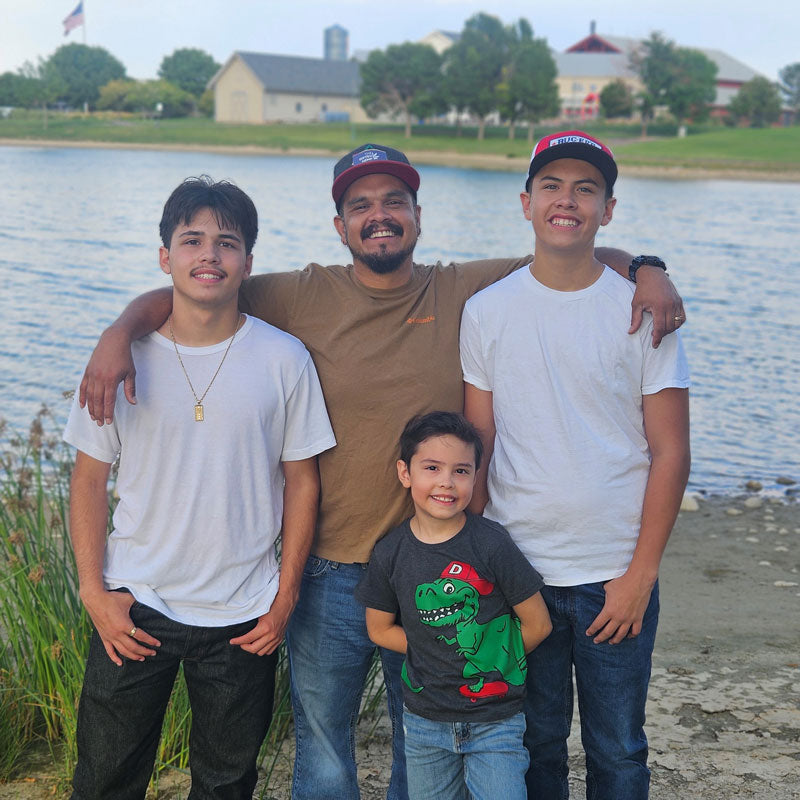 A father with his three sons posing by a pond. The father, in a brown shirt and cap, has his arms around his two older sons, while the youngest son stands in front. The background includes water and distant buildings.