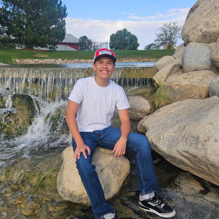 A young boy sitting on a large rock in front of a small waterfall. He is wearing a white shirt, jeans, and a cap, smiling brightly while enjoying the natural setting.