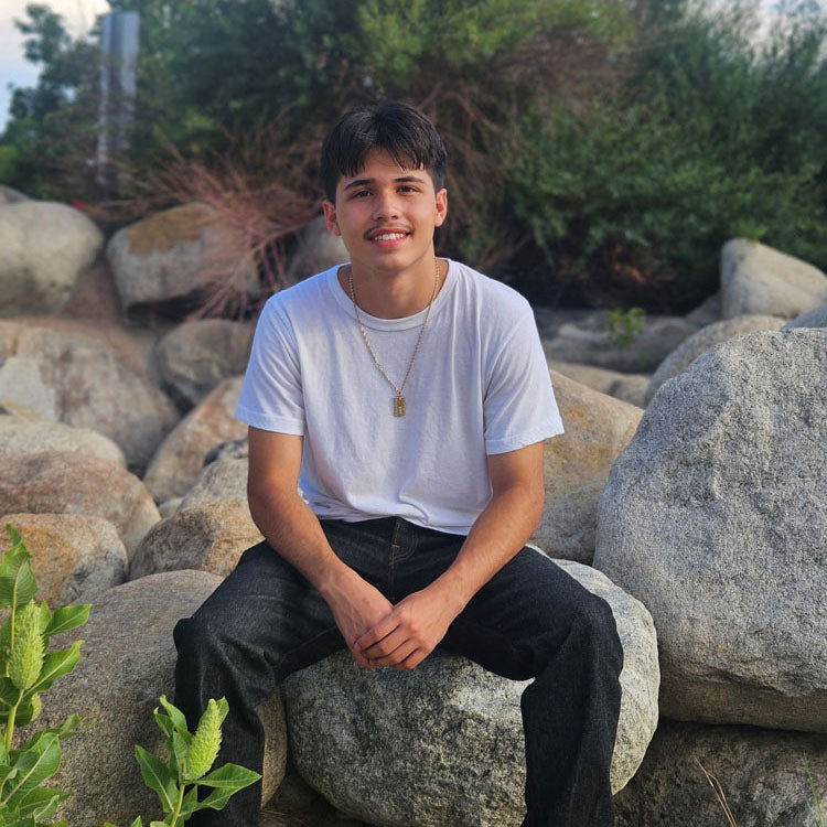 A young man in a white shirt and jeans sitting on large rocks outdoors, smiling at the camera. The background is filled with greenery and boulders.