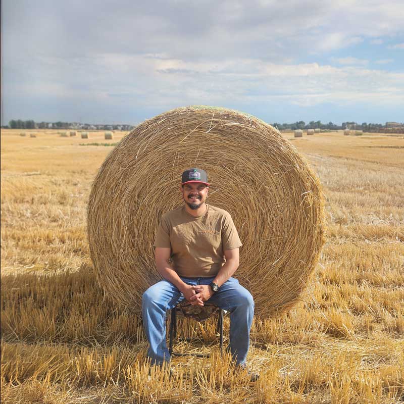 A man sitting on a stool in front of a large hay bale in a harvested field. He is wearing a brown shirt, jeans, and a cap, smiling with the golden landscape of hay bales behind him.