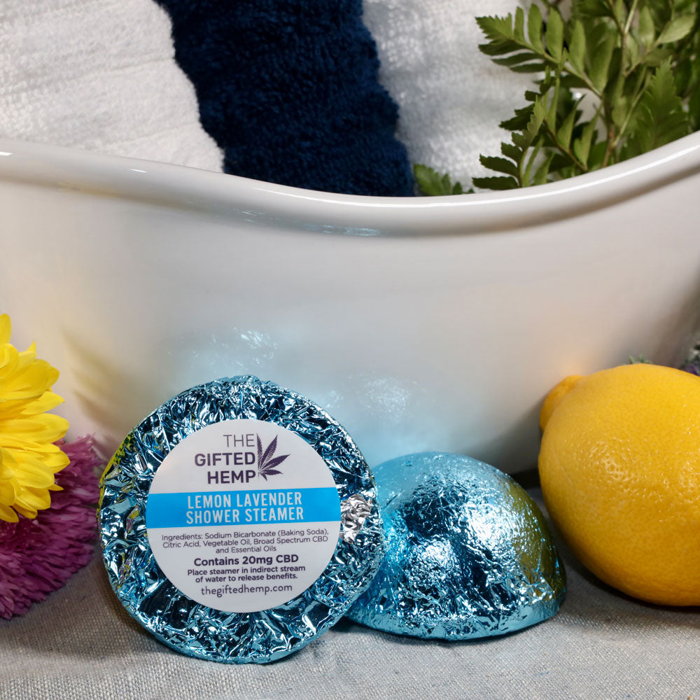 Two Lemon Lavender Shower Steamers from The Gifted Hemp, placed in front of a bath setup with a towel, lemons, and flowers, wrapped in light blue foil.