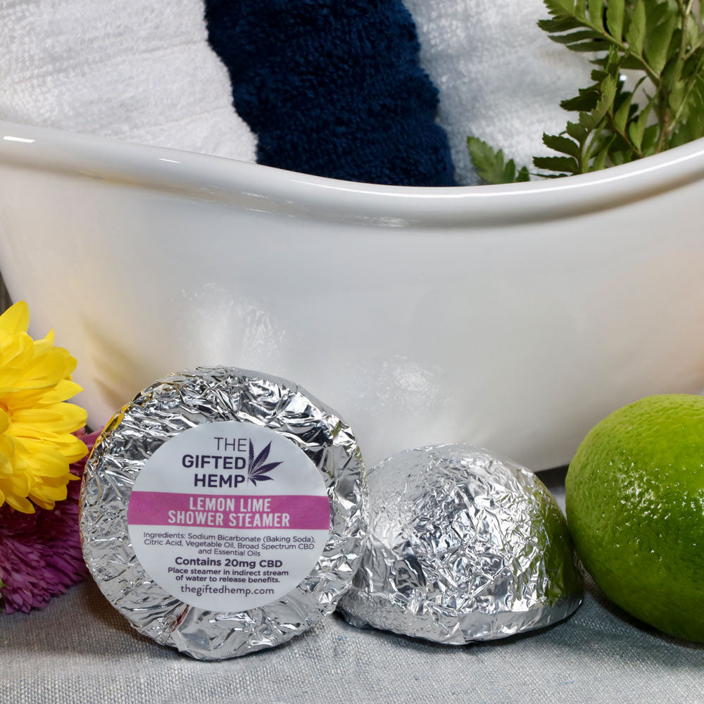 Two Lemon Lime Shower Steamers from The Gifted Hemp, placed in front of a bath setup with a towel, lime, and flowers, wrapped in silver foil.