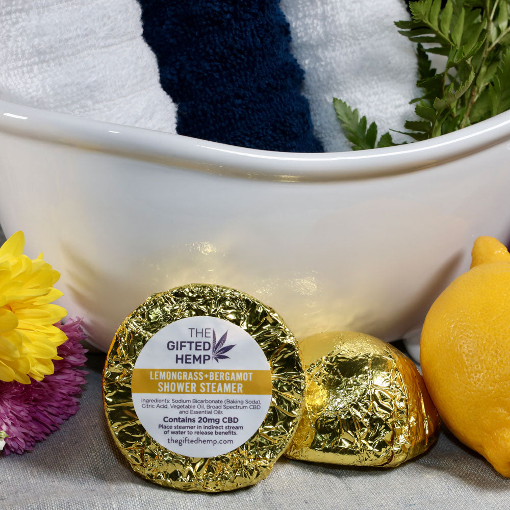 Two Lemongrass Bergamont Shower Steamers from The Gifted Hemp, placed in front of a bath setup with a towel, lemons, and flowers, wrapped in yellow foil.