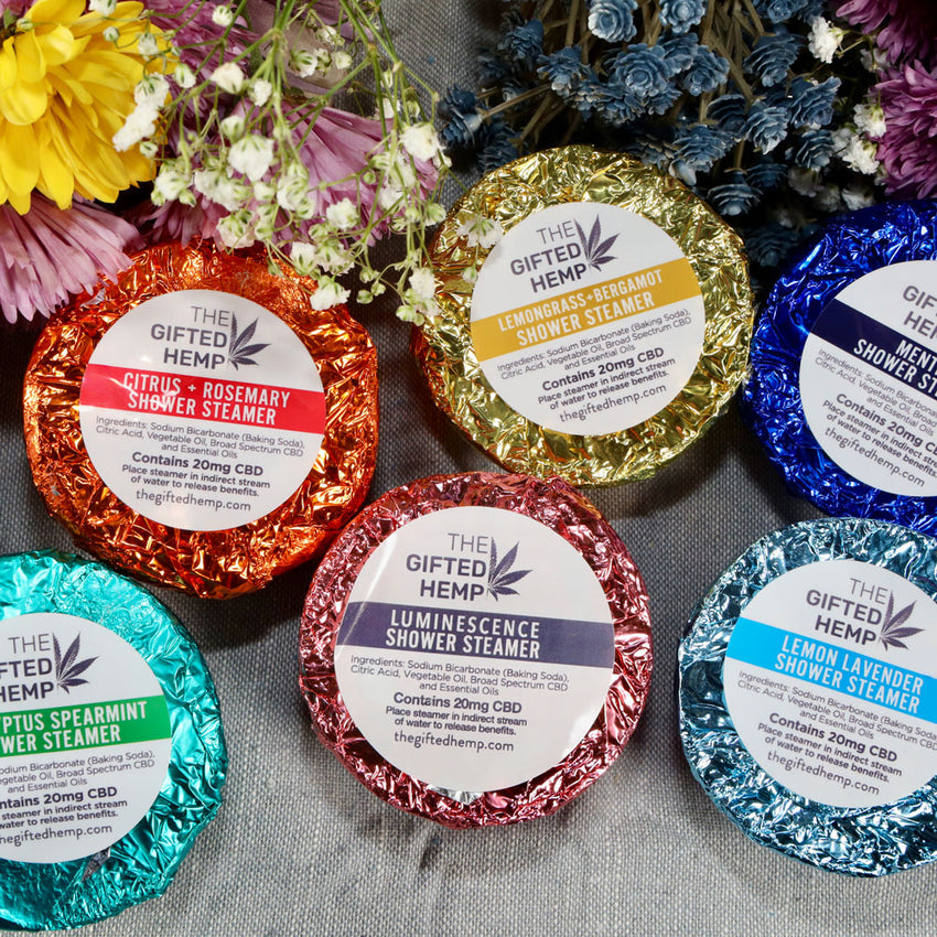 Six colorful wrapped shower steamers from The Gifted Hemp in various scents displayed with fresh flowers